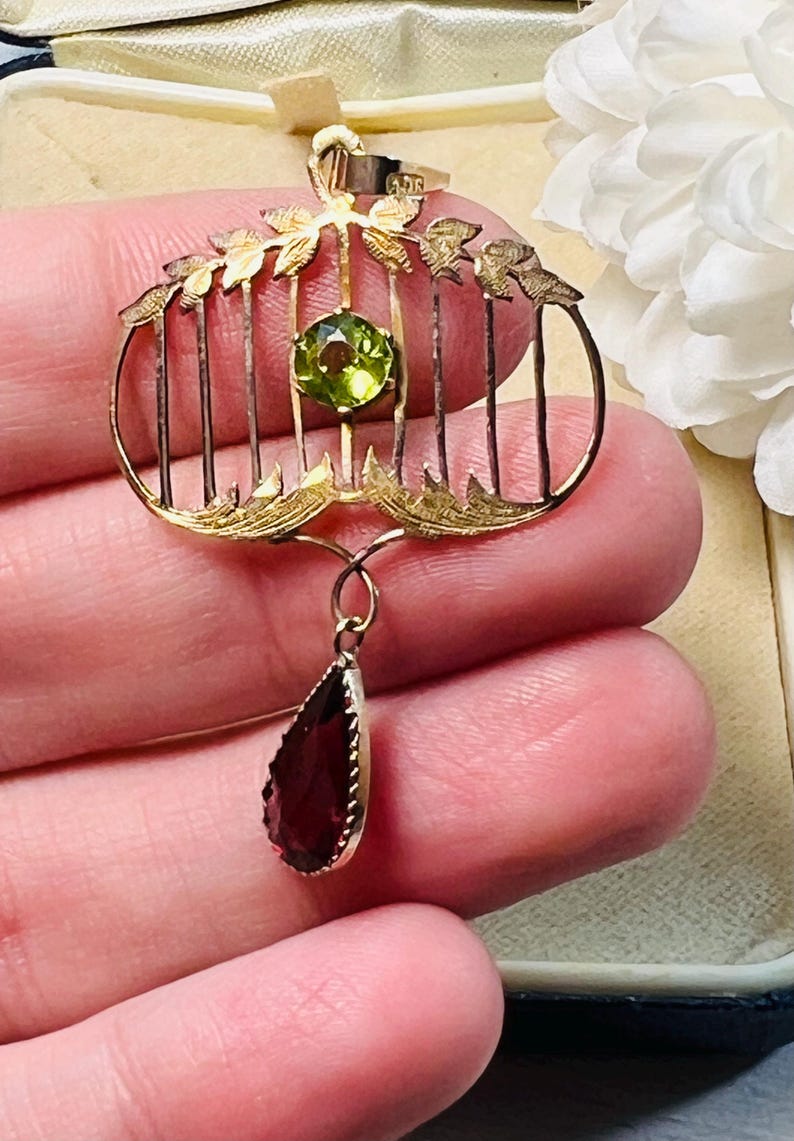 May include: A gold-toned pendant with a cage-like design, featuring a round green gemstone and a teardrop-shaped, dark red gemstone. The pendant has leaf-like accents and vertical bars, creating a vintage aesthetic. The gemstones are the focal point of this jewelry piece.