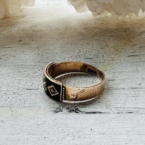 May include: A gold-toned ring with a black enamel band featuring small diamond-shaped accents. The ring is set against a light-colored wooden surface, with a blurred floral element in the background. The ring's design suggests a vintage or antique style.