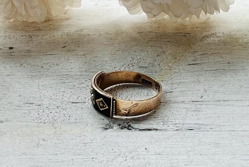 May include: A gold-toned ring with a black enamel band featuring small diamond-shaped accents. The ring is set against a light-colored wooden surface, with a blurred floral element in the background. The ring's design suggests a vintage or antique style.