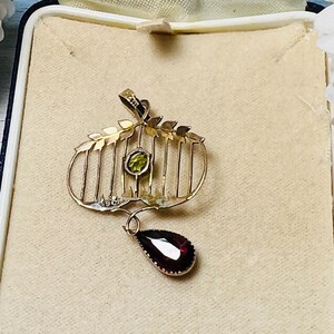 May include: A gold-toned pendant with a cage-like design, featuring a round green gemstone and a teardrop-shaped, deep red gemstone. The pendant is set in a cream-colored box, suggesting a vintage or antique piece.