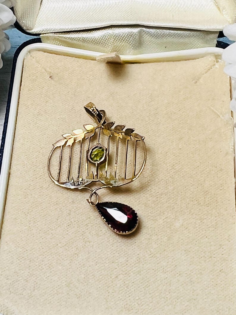 May include: A gold-toned pendant with a cage-like design, featuring a round green gemstone and a teardrop-shaped, deep red gemstone. The pendant is set in a cream-colored box, suggesting a vintage or antique piece.