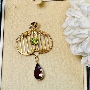 May include: A gold-toned pendant with a cage-like design, featuring a round green gemstone and a teardrop-shaped, dark red gemstone. The pendant is displayed in a vintage jewelry box with a beige interior.