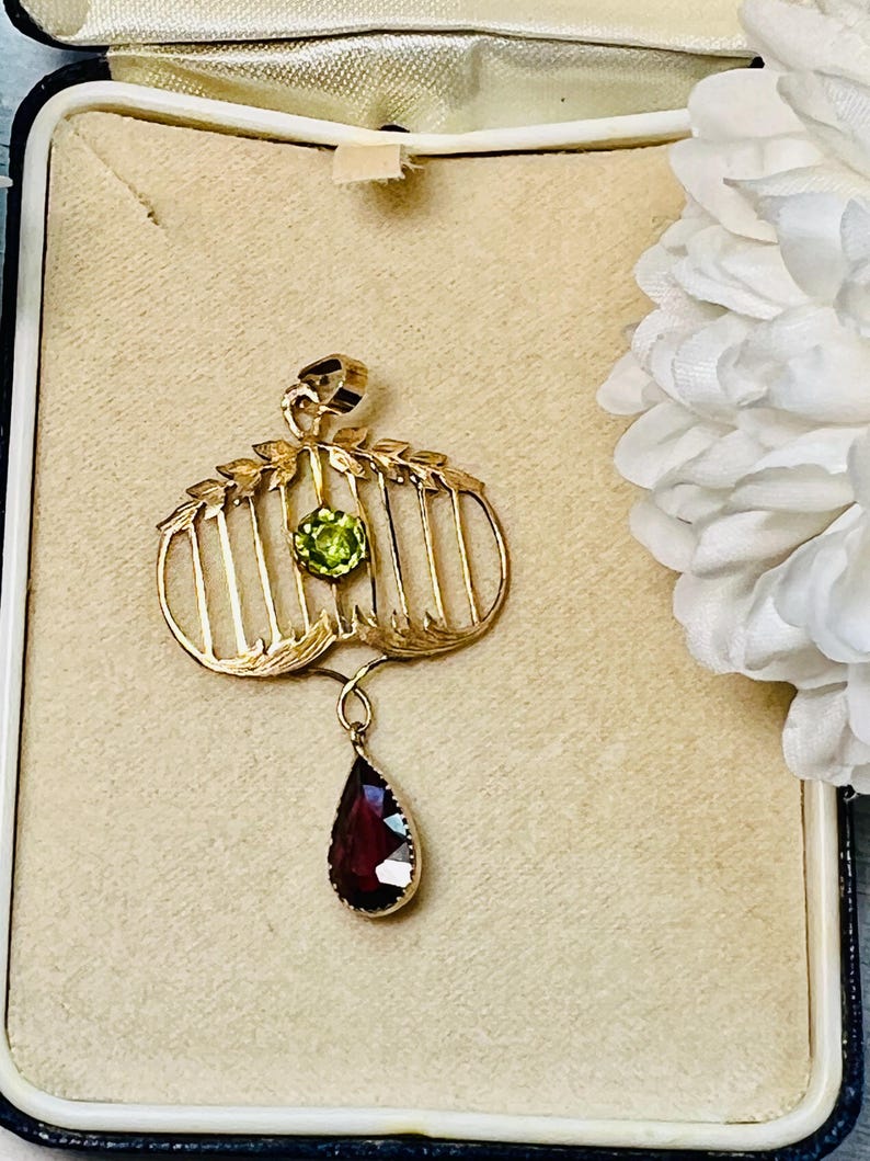 May include: A gold-toned pendant with a cage-like design, featuring a round green gemstone and a teardrop-shaped, dark red gemstone. The pendant is displayed in a vintage jewelry box with a beige interior.
