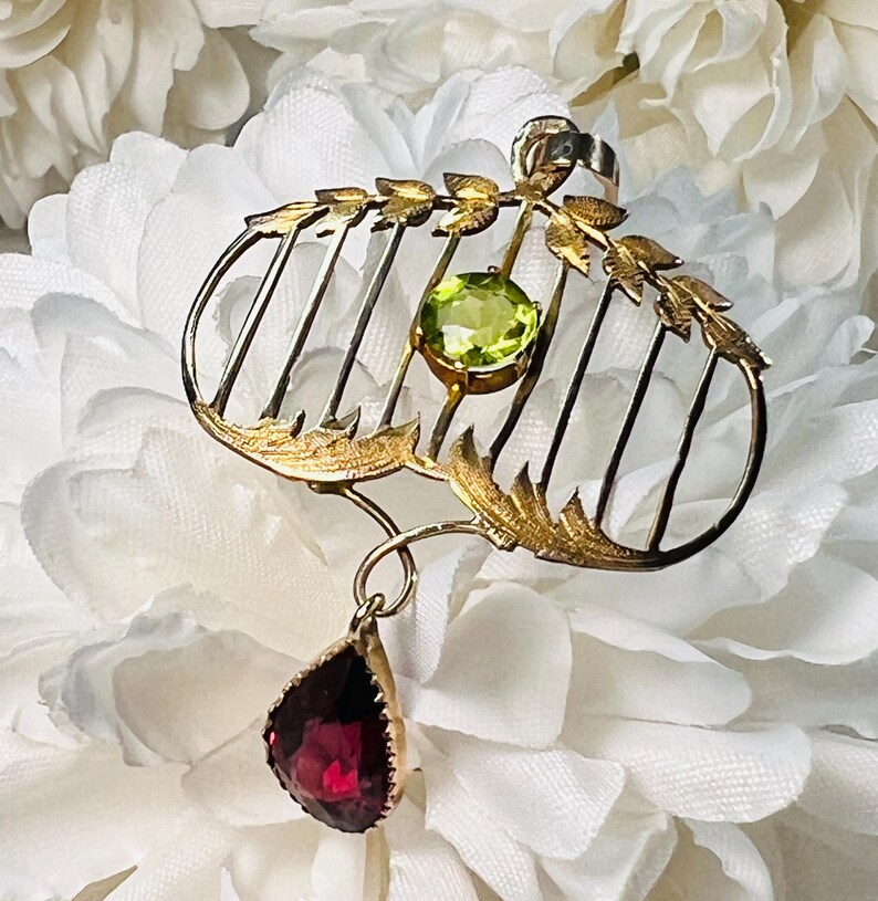 May include: A gold-toned brooch with a cage-like design, featuring a round, light green gemstone and a teardrop-shaped, deep red gemstone pendant. The brooch has leaf-like accents and is set against a white floral background.