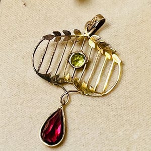May include: A gold-toned pendant with a leaf and cage design, featuring a round green gemstone and a teardrop-shaped red gemstone. The pendant has a delicate, vintage aesthetic, suitable for necklaces.