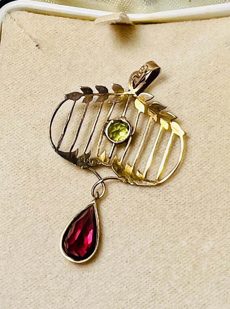 May include: A gold-toned pendant with a leaf and cage design, featuring a round green gemstone and a teardrop-shaped red gemstone. The pendant has a delicate, vintage aesthetic, suitable for necklaces.
