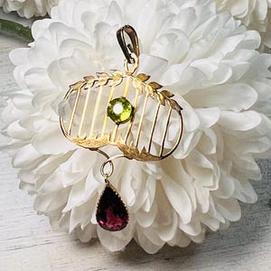 May include: Gold-toned pendant with a cage-like design, featuring a round green gemstone and a teardrop-shaped, deep red gemstone. The pendant has a bail for hanging and is set against a white floral background.