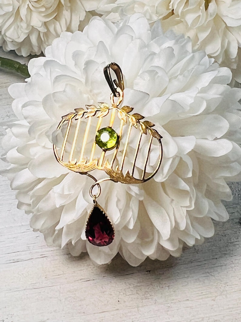 May include: Gold-toned pendant with a cage-like design, featuring a round green gemstone and a teardrop-shaped, deep red gemstone. The pendant has a bail for hanging and is set against a white floral background.