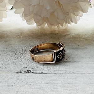 May include: A gold ring with a black enamel band and small diamond accents. The ring is set against a light-colored wooden surface, with white flowers in the background. The ring's design is vintage.