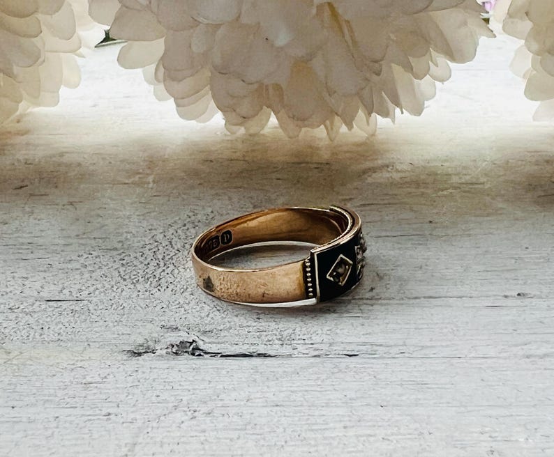 May include: A gold ring with a black enamel band and small diamond accents. The ring is set against a light-colored wooden surface, with white flowers in the background. The ring's design is vintage.