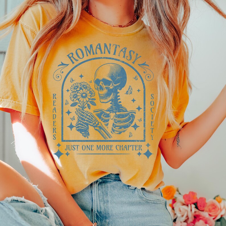 May include: A yellow t-shirt with a blue graphic design featuring a skeleton holding flowers. The graphic also includes the text "Romantasy Readers Society" and "Just One More Chapter".