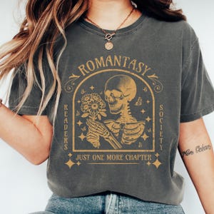 May include: A dark gray t-shirt with a vintage-style graphic of a skeleton holding a bouquet of flowers. The graphic is surrounded by a banner that reads "Romantasy Readers Society" and "Just One More Chapter".