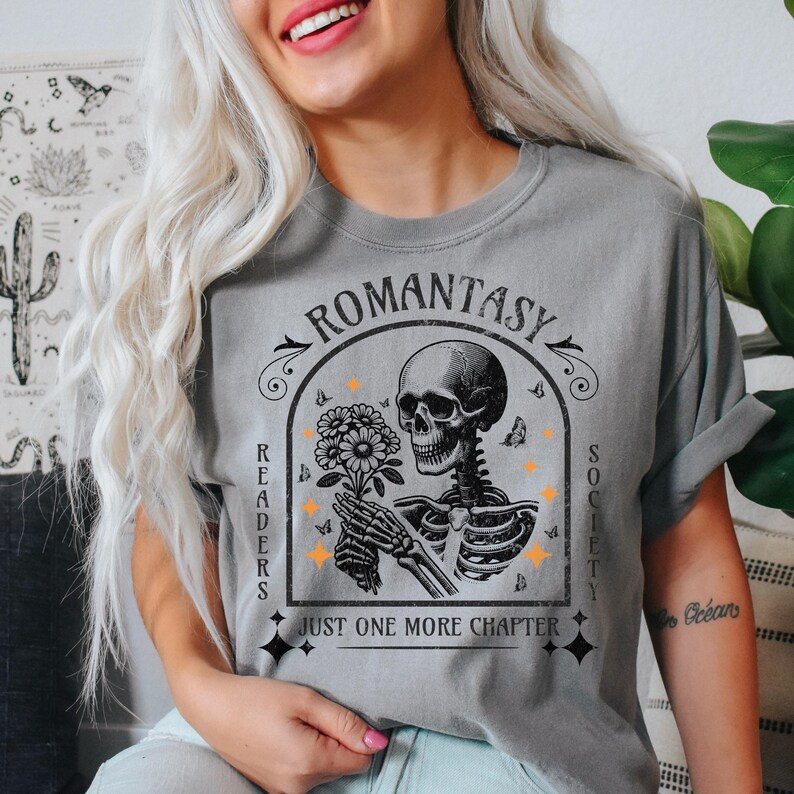 May include: Gray t-shirt with a black and white illustration of a skeleton holding a flower. The text "ROMANTASY READERS SOCIETY JUST ONE MORE CHAPTER" is printed on the shirt.
