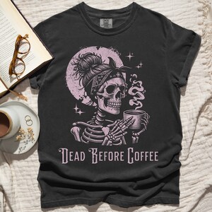 May include: Black t-shirt with a pink and white illustration of a skeleton holding a coffee cup. The skeleton has a bandana on its head and is surrounded by stars. The text "Dead Before Coffee" is printed below the illustration.