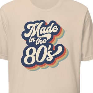 Puede incluir: Camiseta color crema con el texto "Made in the 80's" en una fuente retro. El texto es blanco con un contorno azul oscuro y un efecto de sombra colorido en capas. La camiseta tiene mangas cortas.