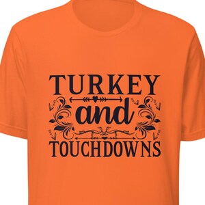 May include: An orange t-shirt featuring the text "TURKEY and TOUCHDOWNS" in black, decorative lettering. The shirt has a crew neck and is a solid orange colour.