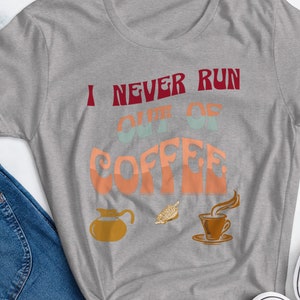 Op de afbeelding: Een grijze t-shirt met de tekst "I NEVER RUN OUT OF COFFEE" in retro kleuren. Het ontwerp bevat koffie-gerelateerde afbeeldingen: een koffiepot, bonen en een kopje. Het shirt wordt gecombineerd met een blauwe jeans en zwarte sneakers.