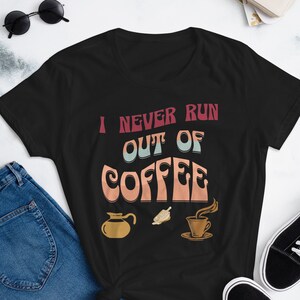 May include: Black t-shirt with the text "I NEVER RUN OUT OF COFFEE" in a retro font. The shirt features coffee-related graphics, including a coffee pot, a coffee bean, and a cup of coffee. The shirt is paired with blue jeans and black trainers.
