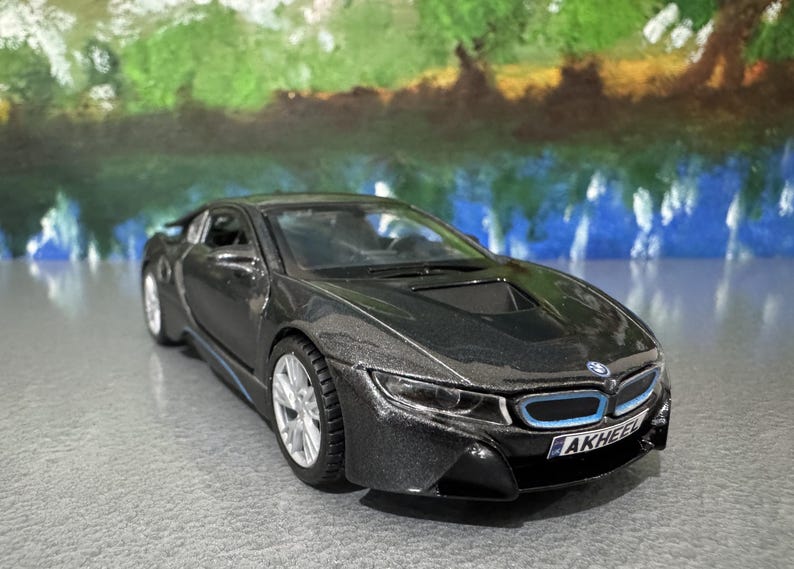 May include: A dark gray model car with silver wheels and blue accents. The car has a sleek, modern design with a low profile and a prominent front grille. The license plate reads "AKHEEL".