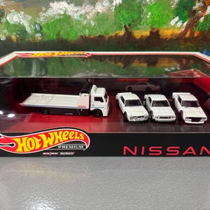 May include: A Hot Wheels Premium Nissan die-cast car set. The set includes a white car carrier truck and three white Nissan cars. The box is black with red accents and the Hot Wheels logo.