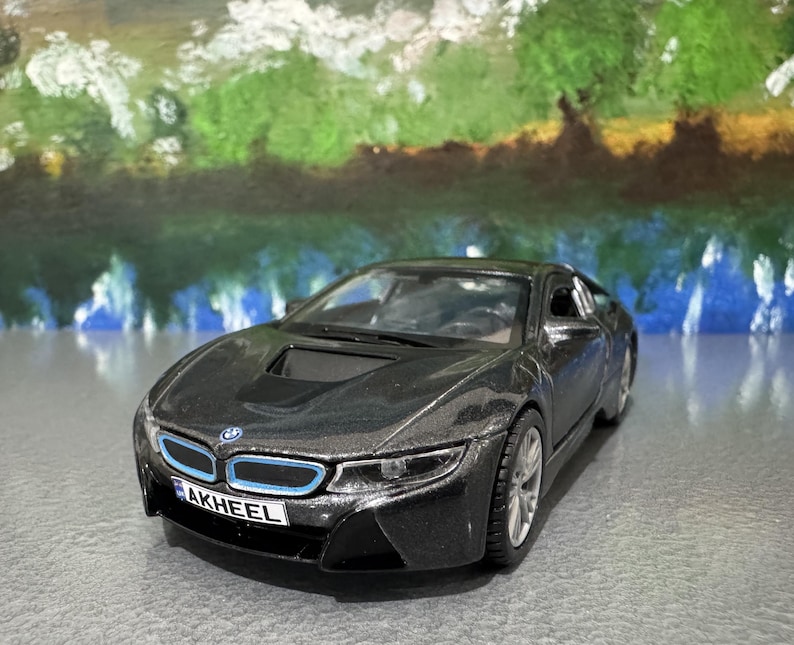 May include: A dark gray BMW i8 model car with blue accents on the front grill. The toy car has a black front bumper and a license plate that reads "AKHEEL". The car is on a gray surface with a blurred background.