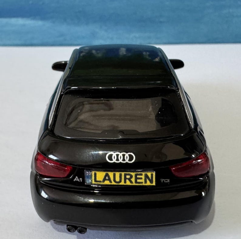 Peut inclure: Une maquette noire d'Audi A1 avec une plaque d'immatriculation jaune portant le nom "LAUREN". La voiture a des feux arri&egrave;re rouges et le logo Audi. R&eacute;plique miniature.