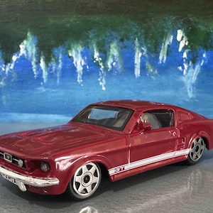 May include: A burgundy toy car with a white stripe and the text "GT" on the side. The car has silver rims and a chrome bumper. The background features a blue and green painted design.