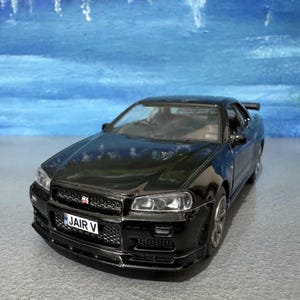May include: A black model car with a spoiler and the license plate "JAIR V". The car has a detailed front grille and headlights. The background is a blurry blue.