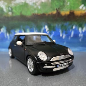 Könnte beinhalten: Ein schwarzes Mini Cooper Spielzeugauto mit weißem Dach. Das Modellauto hat ein Nummernschild mit der Aufschrift "BIG DANG". Das Auto steht auf einer grauen Oberfläche mit verschwommenem Hintergrund.