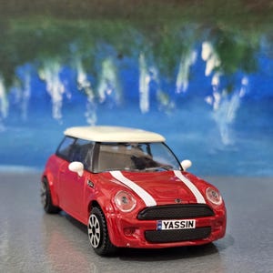 May include: A red toy car with a white roof and white racing stripes. The car has black wheels and the license plate reads "YASSIN". The background features a blue and green abstract painting.