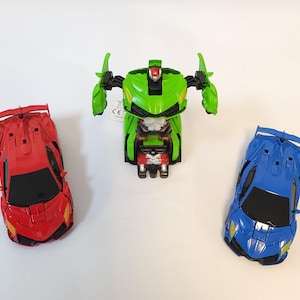May include: Three toy robot cars that transform into robots. One is green, one is red, and one is blue. The green robot is in robot mode, while the red and blue cars are in car mode. The packaging for the toys is visible in the background.
