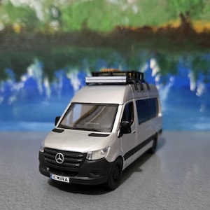 May include: Silver van model with black accents, a roof rack, and the word "EMIRA" on the license plate. The van has a Mercedes-Benz logo on the front and is set against a blurred blue and green background.