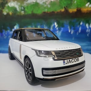 May include: White toy car model of a Range Rover with a gold trim. The car has a black grille and a number plate that reads "JACOB".