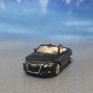 Op de afbeelding: Een zwarte speelgoed cabriolet met een beige interieur. De auto heeft het Audi-logo op de grille en zilveren velgen. De achtergrond is een wazige blauwe lucht.