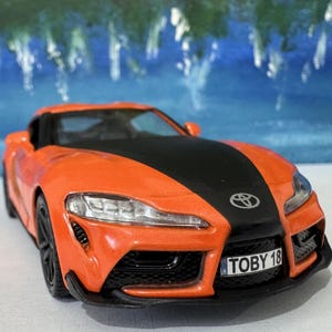 Op de afbeelding: Oranje en zwarte speelgoedauto met een zwarte race streep en de tekst "TOBY 18" op het kenteken.