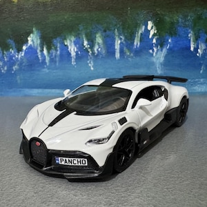 May include: A white and black toy sports car with black wheels and a rear spoiler. The car has the word "PANCHO" on the license plate. The car is on a gray surface with a blue and green background.