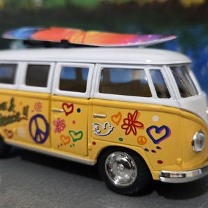 Puede incluir: Una furgoneta de juguete amarilla y blanca con una tabla de surf en el techo. La furgoneta está decorada con símbolos de paz, corazones y flores. Las palabras "love & peace" están escritas en el lateral. Tiene neumáticos negros y tapacubos cromados.