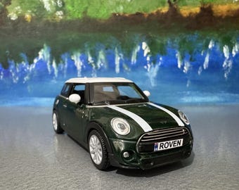 Puede incluir: Un coche de juguete Mini Cooper verde oscuro con techo blanco y franjas blancas. El coche de juguete tiene ruedas plateadas y una matrícula que dice "ROVEN". El fondo es un paisaje pintado.
