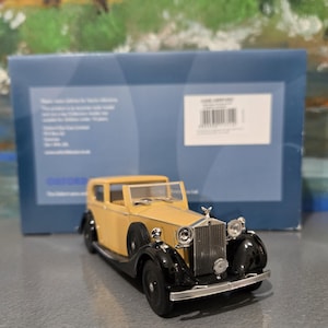 Könnte beinhalten: Gelb-schwarzes Modellauto eines Oldtimer Rolls Royce mit silbernem Frontgrill und Kühlerfigur. Das Modell steht vor einer blauen Schachtel.