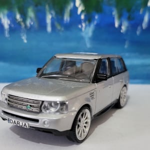 May include: Silver SUV model with black trim and white wheels. The front of the vehicle has a black grill and the license plate reads "DARJA". The background is a blurred blue and green.