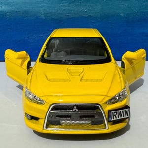 May include: A yellow die-cast toy car model of a Mitsubishi Lancer Evolution with the doors open. The car has a black front grille and a black license plate that reads "IRWIN".