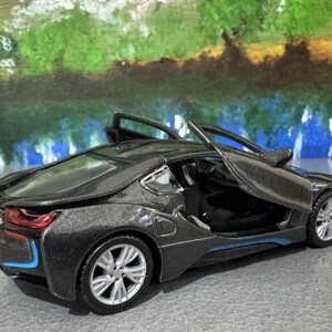May include: A dark gray toy car with its doors open, showcasing a detailed interior. The car has silver rims and blue accents. The background features a colorful, abstract painting.