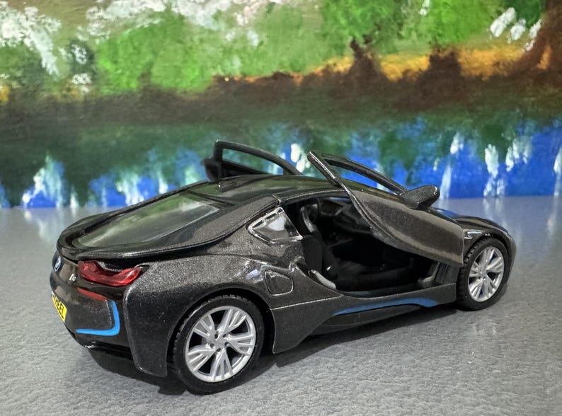 May include: A dark gray toy car with its doors open, showcasing a detailed interior. The car has silver rims and blue accents. The background features a colorful, abstract painting.