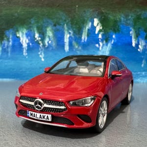 Puede incluir: Un coche modelo Mercedes-Benz CLA rojo con techo negro. El coche tiene una parrilla y faros plateados. La matrícula dice "MALAKA". El fondo es una pintura abstracta azul y verde.