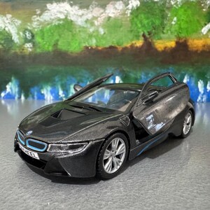 May include: A dark gray toy car with the doors open, showcasing a sleek design. The car has silver wheels and blue accents. The background features a painted landscape with water and trees.