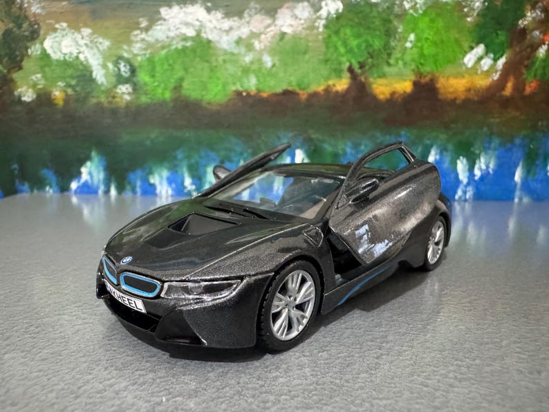 May include: A dark gray toy car with the doors open, showcasing a sleek design. The car has silver wheels and blue accents. The background features a painted landscape with water and trees.