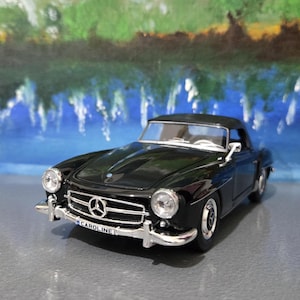 Może przedstawiać: Czarny model zabytkowego samochodu z otwieranym dachem z chromowanymi detalami. Samochód ma czarny dach i srebrną przednią kratkę z logo Mercedes-Benz. Tablica rejestracyjna brzmi "CAROLINE". Tło to rozmyty obraz krajobrazu.