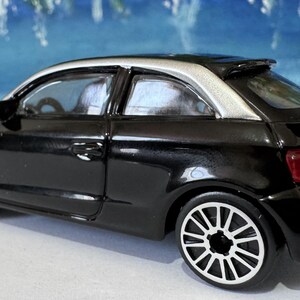 Peut inclure: Une petite voiture noire avec un toit argent&eacute; et des jantes blanches. Le v&eacute;hicule miniature a un aileron et des feux arri&egrave;re rouges. C'est un mod&egrave;le r&eacute;duit, probablement un jouet ou un objet de collection.