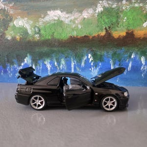 Op de afbeelding: Een zwarte speelgoedauto met de motorkap, kofferbak en deur open, waardoor het interieur zichtbaar is. De auto heeft zilveren velgen en staat tegen een achtergrond van een geschilderd landschap met blauwe, groene en bruine kleuren.