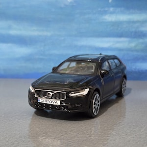 May include: A black Volvo estate car model with the number plate "CHITOVA". The miniature car has a detailed grill, headlights, and wheels. The background is a gradient of blue and white.